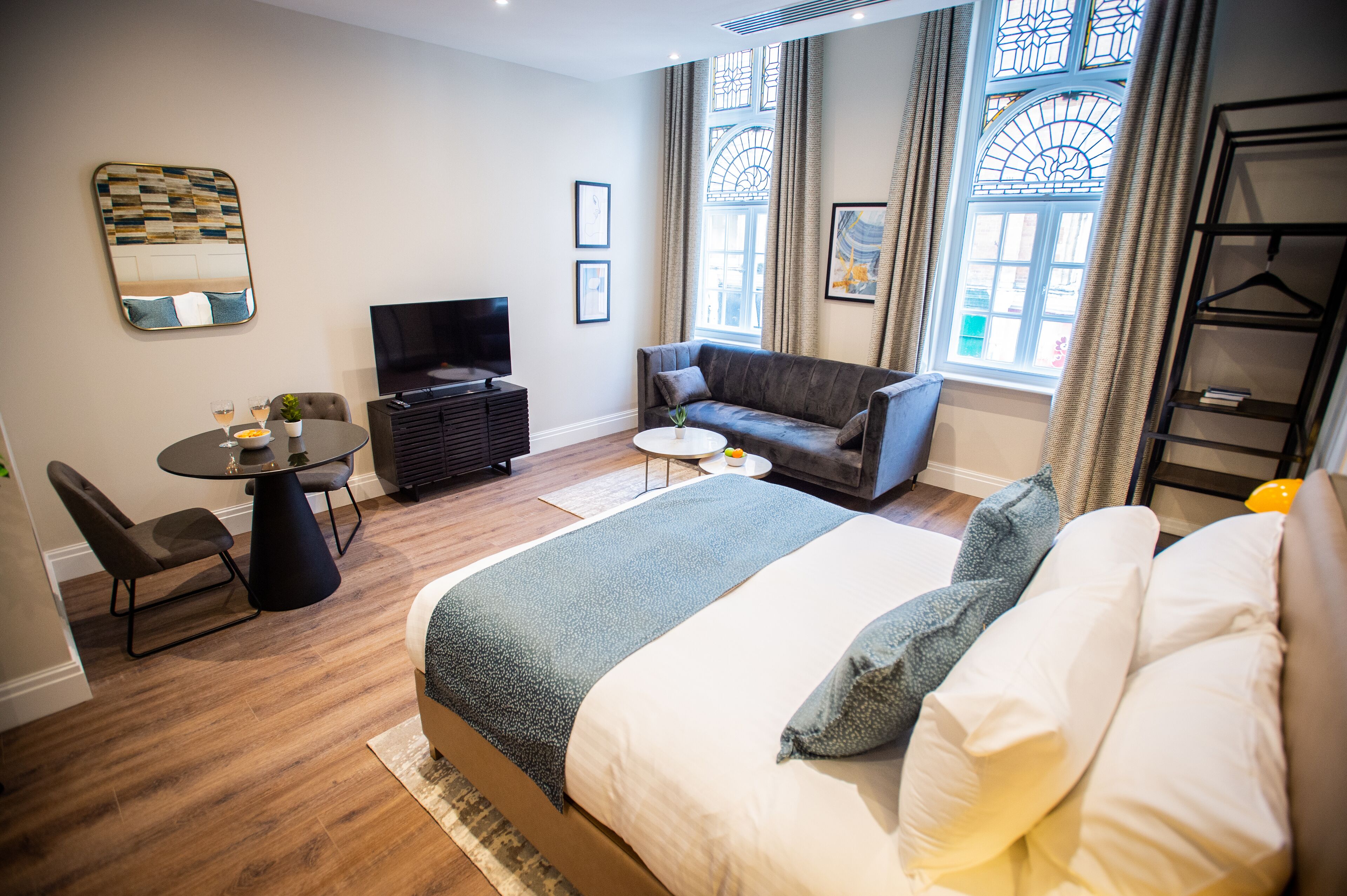 A modern hotel room with a large bed, a sofa, a TV, a dining table, and a small balcony.