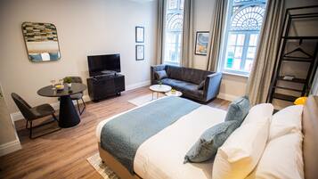 A modern hotel room with a large bed, a sofa, a TV, a dining table, and a small balcony.
