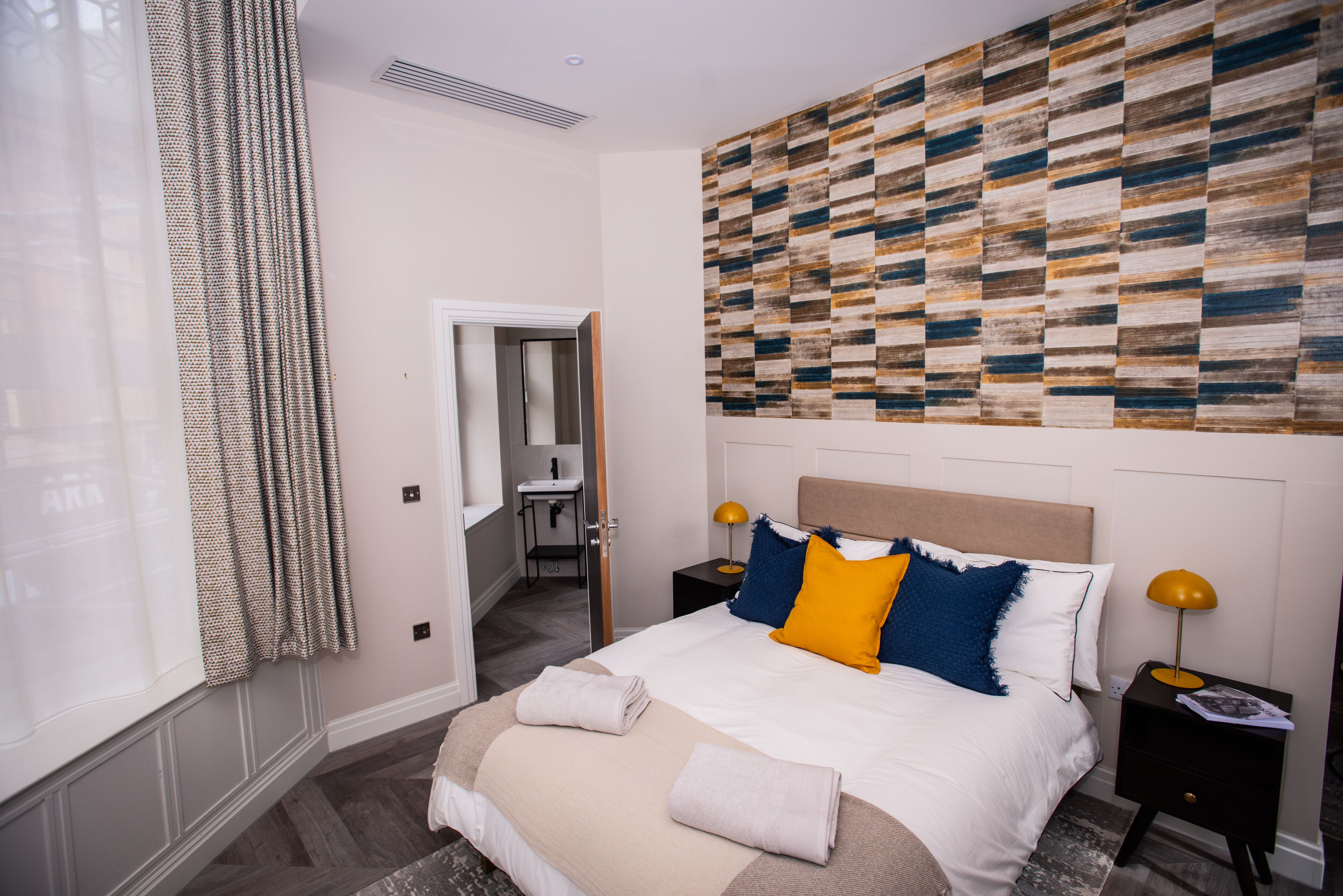 Habitació d'hotel amb un llit, una tauleta de nit, una obra d'art a la paret i un bany visible a través d'una porta oberta.