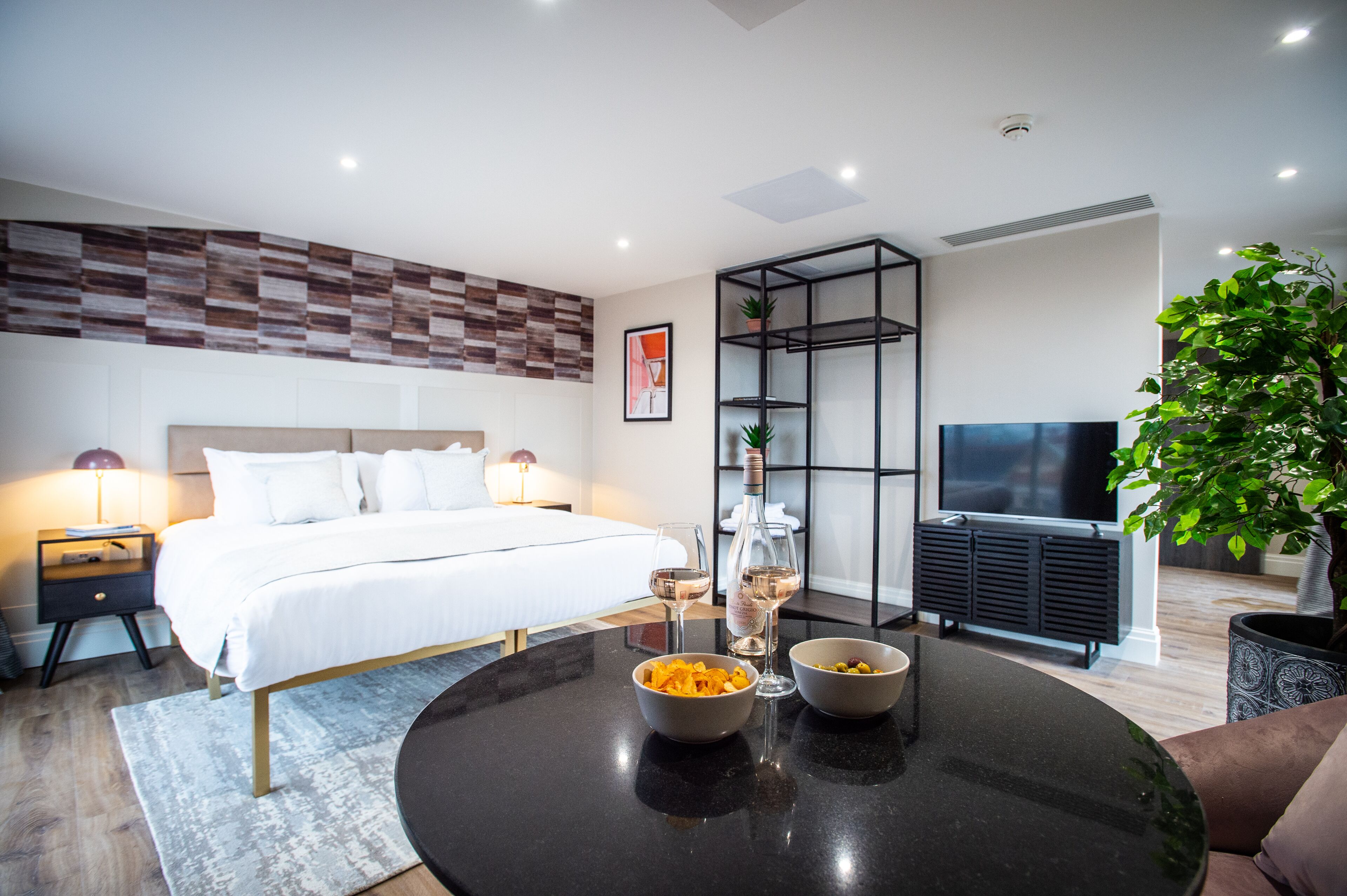 A modern hotel room with a bed, bedside tables, a TV, a glass cabinet, and a dining table with bowls.