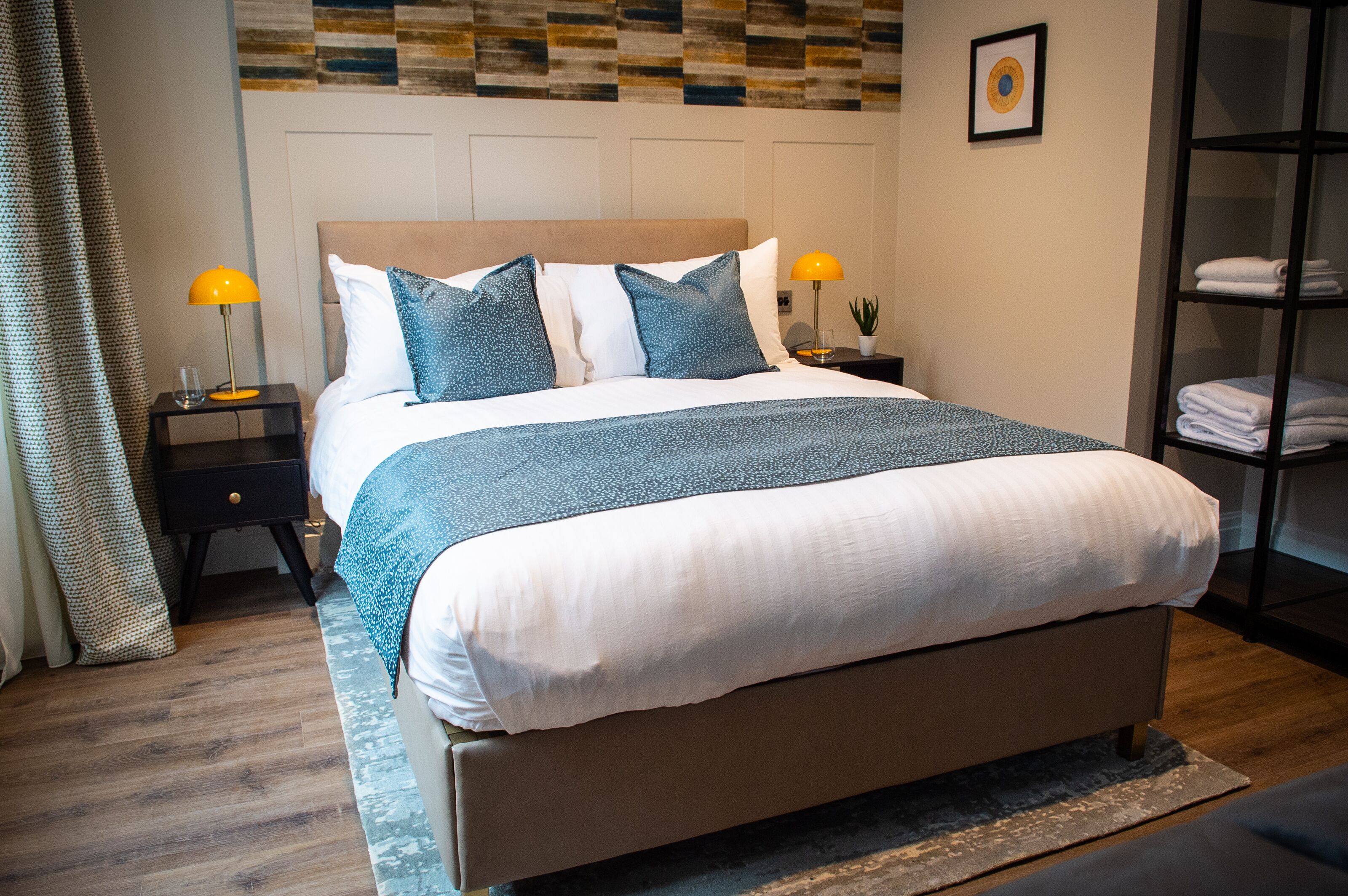 A neatly made bed with a patterned bedspread, two bedside lamps, a nightstand, and a wall-mounted shelf with folded towels.