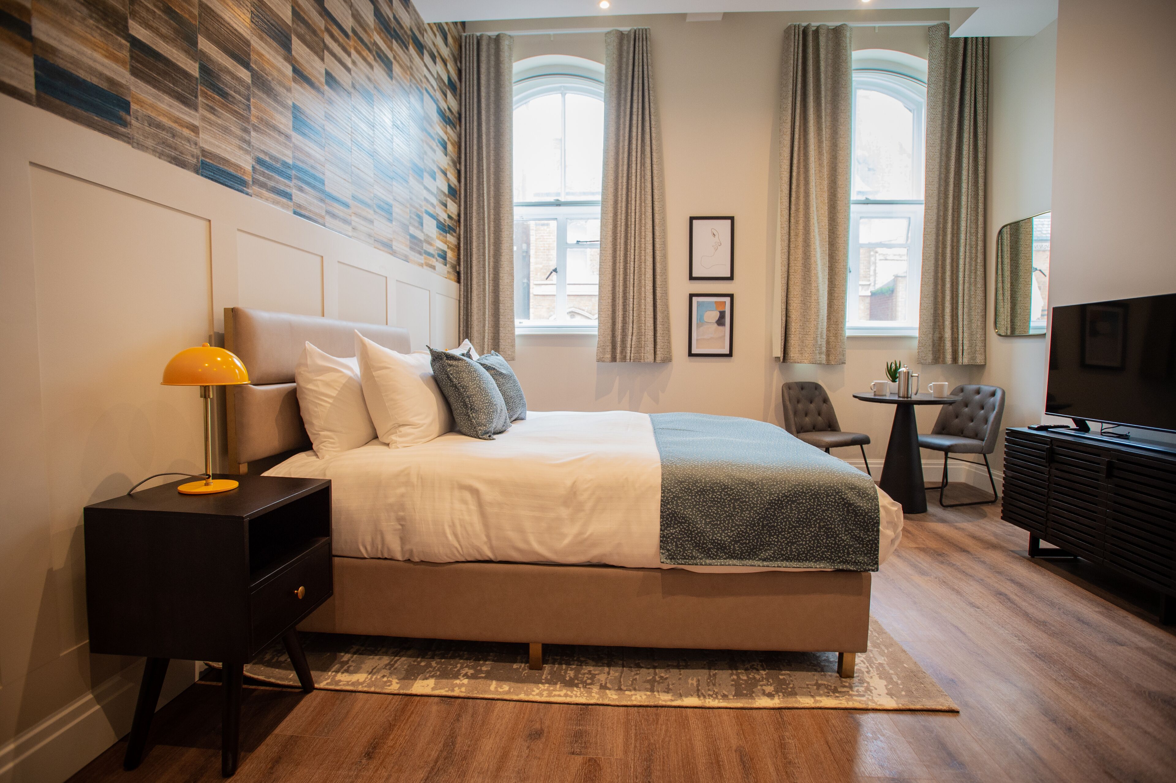 Una habitación de hotel moderna con una cama grande, una mesita de noche con lámpara, un escritorio pequeño con silla, un televisor y vistas a la ciudad a través de amplios ventanales.