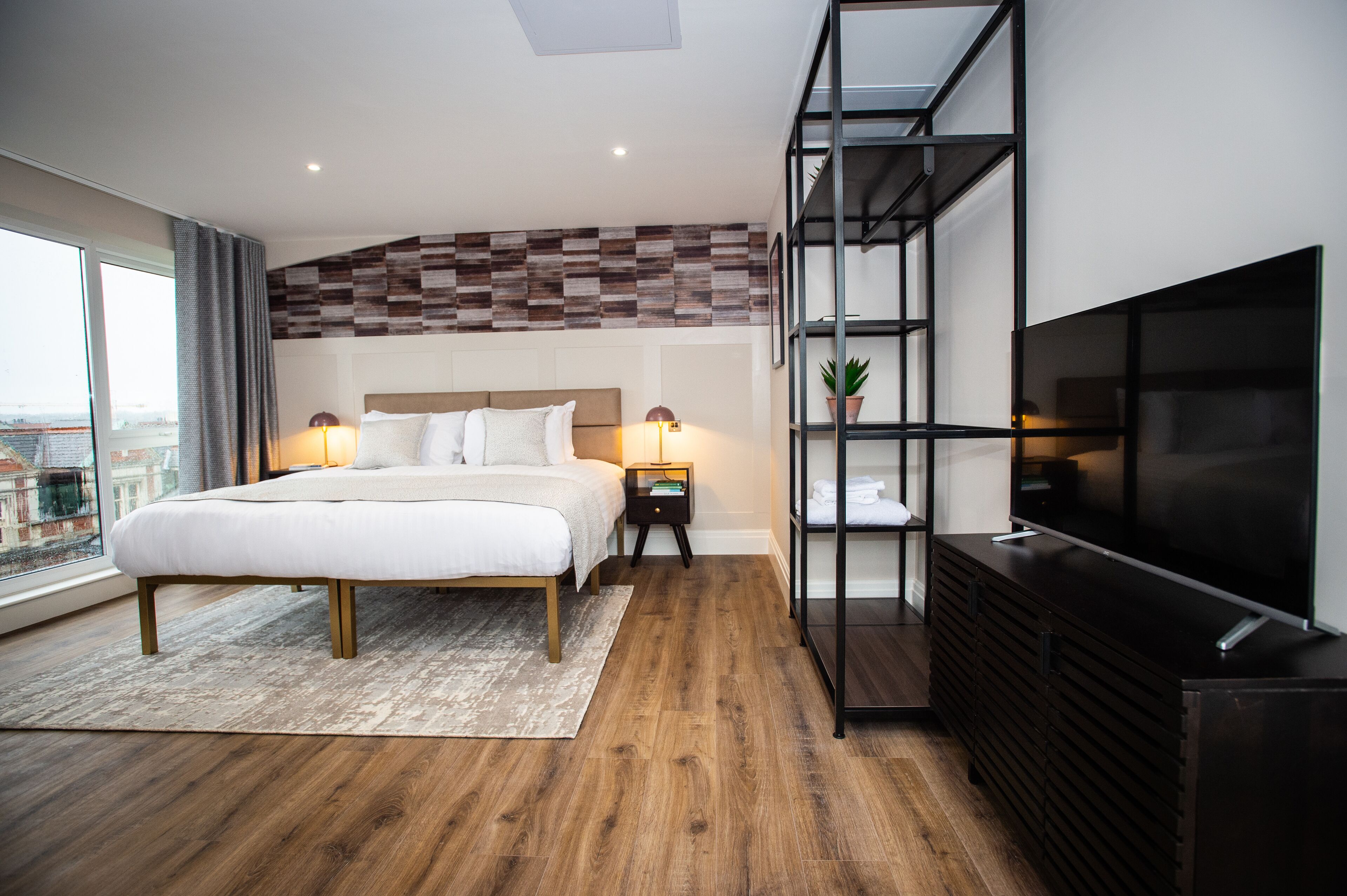 A modern hotel room with a large bed, a TV, a wooden floor, and a city view through the window.