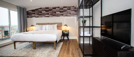 A modern hotel room with a large bed, a TV, a wooden floor, and a city view through the window.