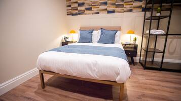 A neatly made bed with a wooden frame and white bedding, a wall with a geometric patterned design, and a metal shelving unit with towels.