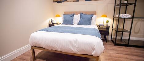 Una cama bien tendida con estructura de madera y ropa de cama blanca, una pared con diseño geométrico y un estante metálico con toallas.