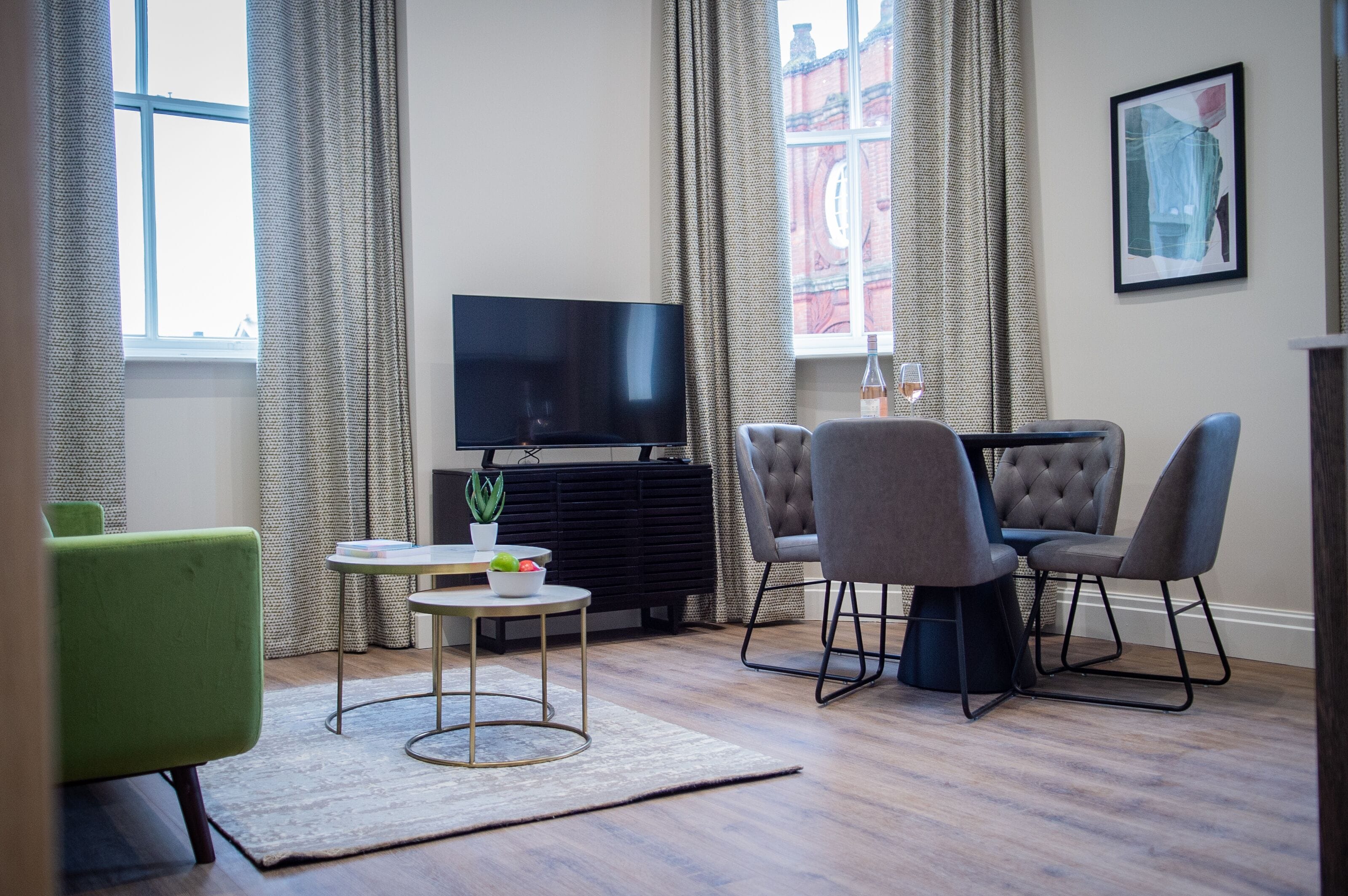 Een moderne woonkamer met een flatscreen televisie, een eettafel met vier stoelen, een klein bijzettafeltje met een plant en een groot raam met gordijnen.