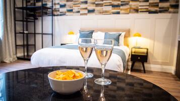 A hotel room with a bed, two bedside lamps, a black table with a bowl of snacks, and two glasses of champagne.