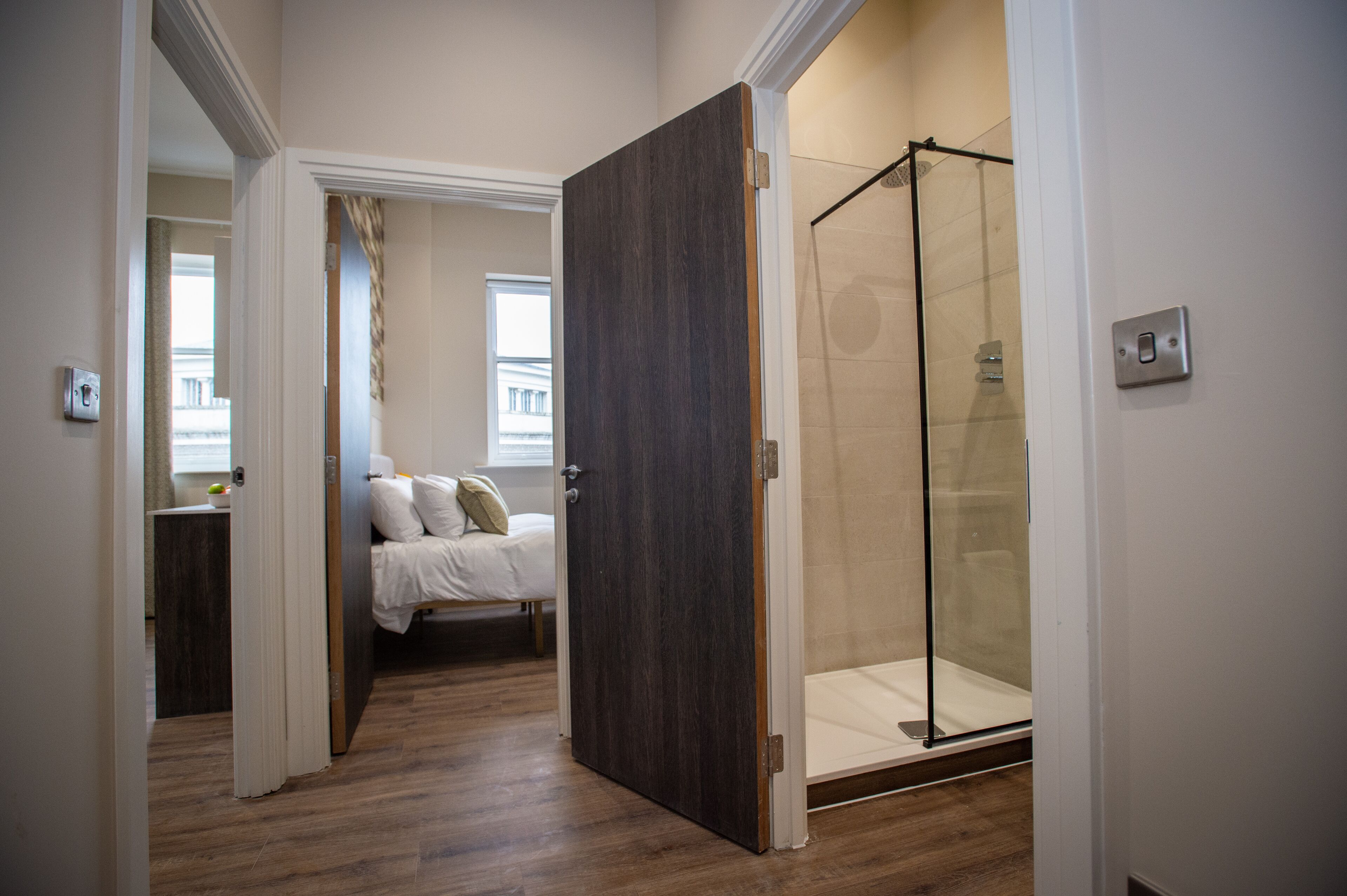 Ein Hotelzimmer mit Holzboden, einem Bett mit weißer Bettwäsche, einer Glasdusche und Blick auf ein Gebäude durch ein Fenster.