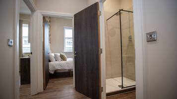 Ein Hotelzimmer mit Holzboden, einem Bett mit weißer Bettwäsche, einer Glasdusche und Blick auf ein Gebäude durch ein Fenster.