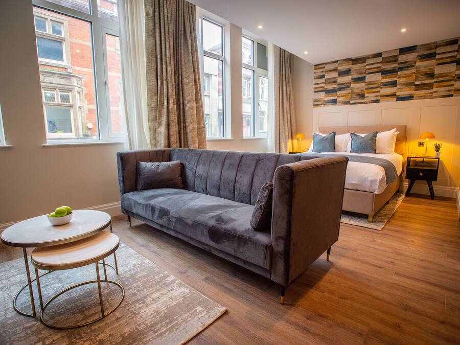 A modern hotel room with a grey sofa, a small round table with a bowl of fruit, a bed with a white headboard, and a view of a brick building through large windows.