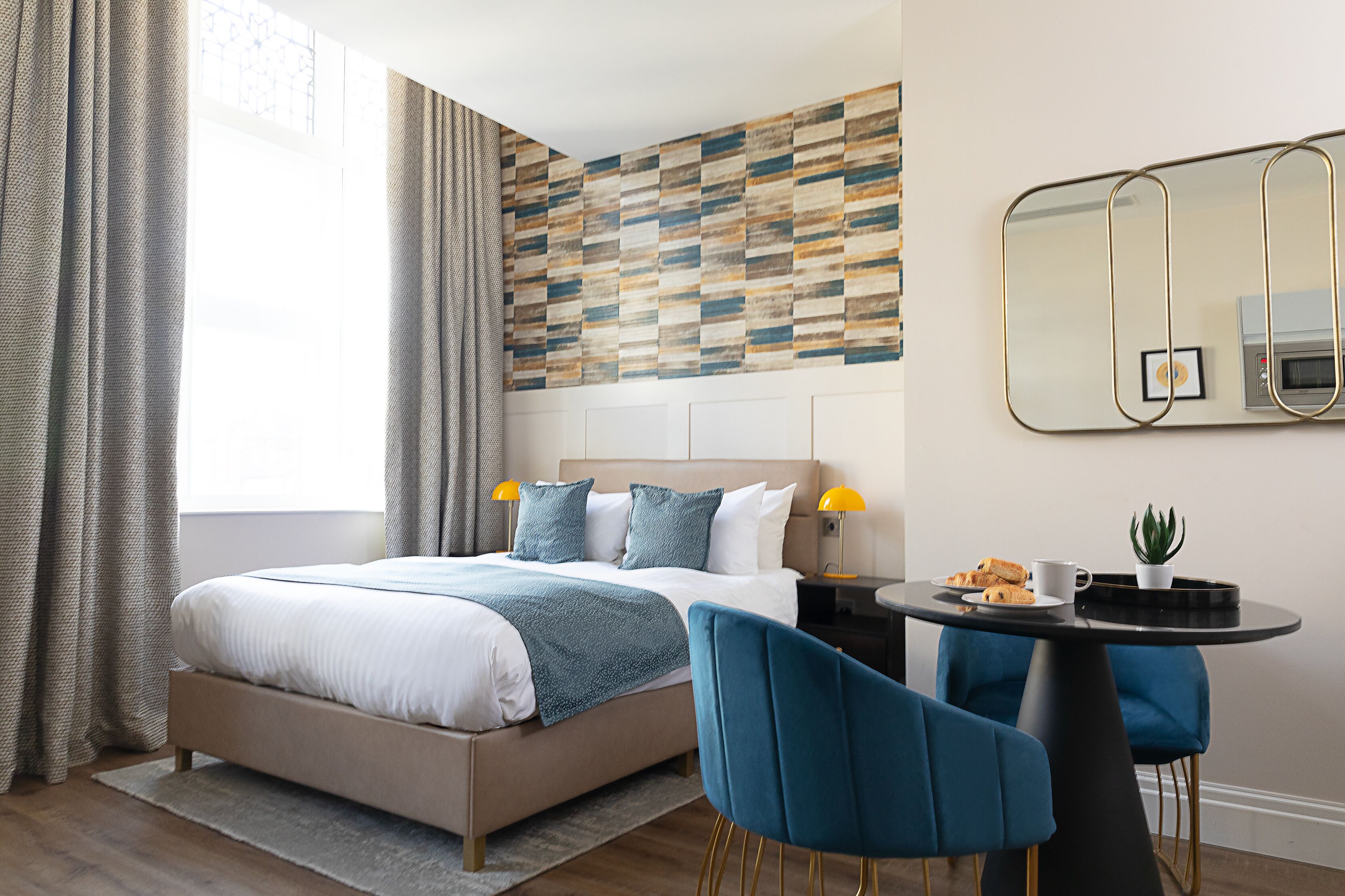 A modern hotel room with a large bed, a bedside table with a lamp, a small round table with a chair, and a decorative wall panel.