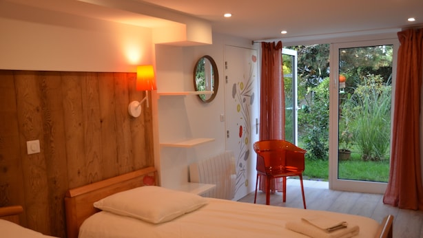 Quarto de hotel com duas camas, cabeceira de madeira, uma cadeira vermelha e uma janela grande com vista para áreas verdes.