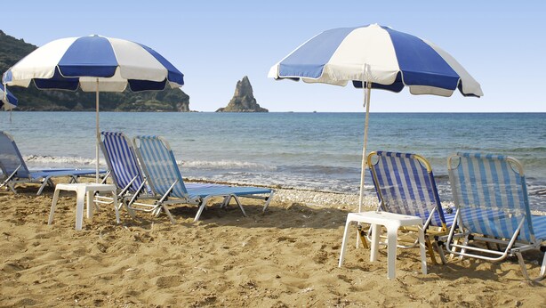 Una playa con varias sillas y sombrillas a rayas azules y blancas, un promontorio rocoso a lo lejos, y un mar tranquilo.