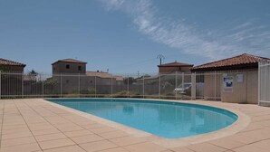 Una piscina con solárium empedrado, rodeada por una valla blanca, y edificios con tejados rojos al fondo.
