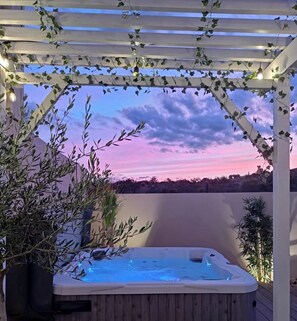 A hot tub under a pergola with ivy, surrounded by plants, and a logo for "Nuit D'Ete Hébergement".