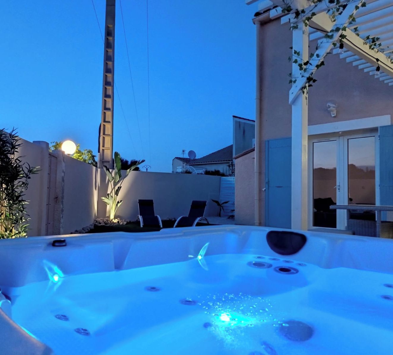 Un bain à remous avec un éclairage bleu, entouré d’un mur blanc et d’une pergola couverte de plantes grimpantes.