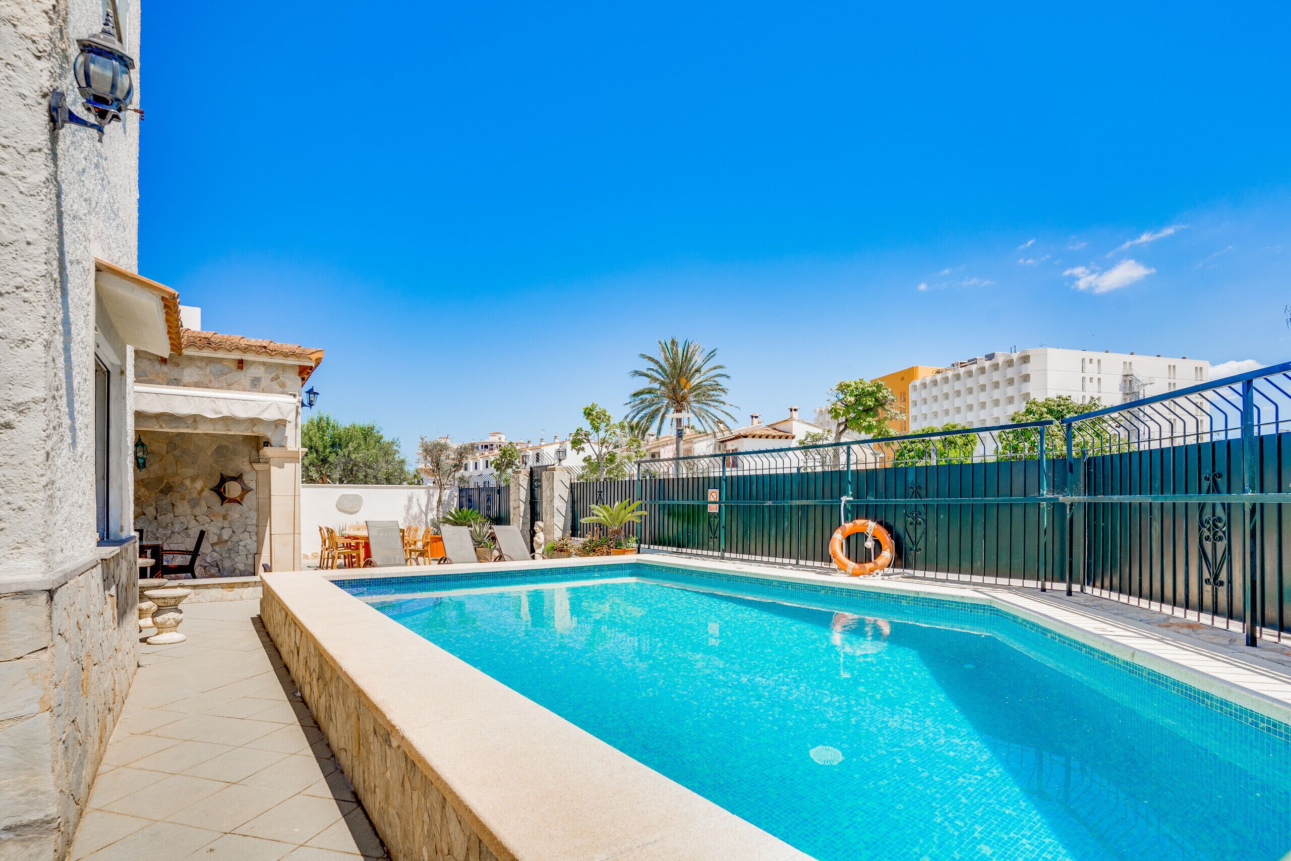 A swimming pool with a stone wall and a tiled deck, surrounded by a fence and a palm tree.