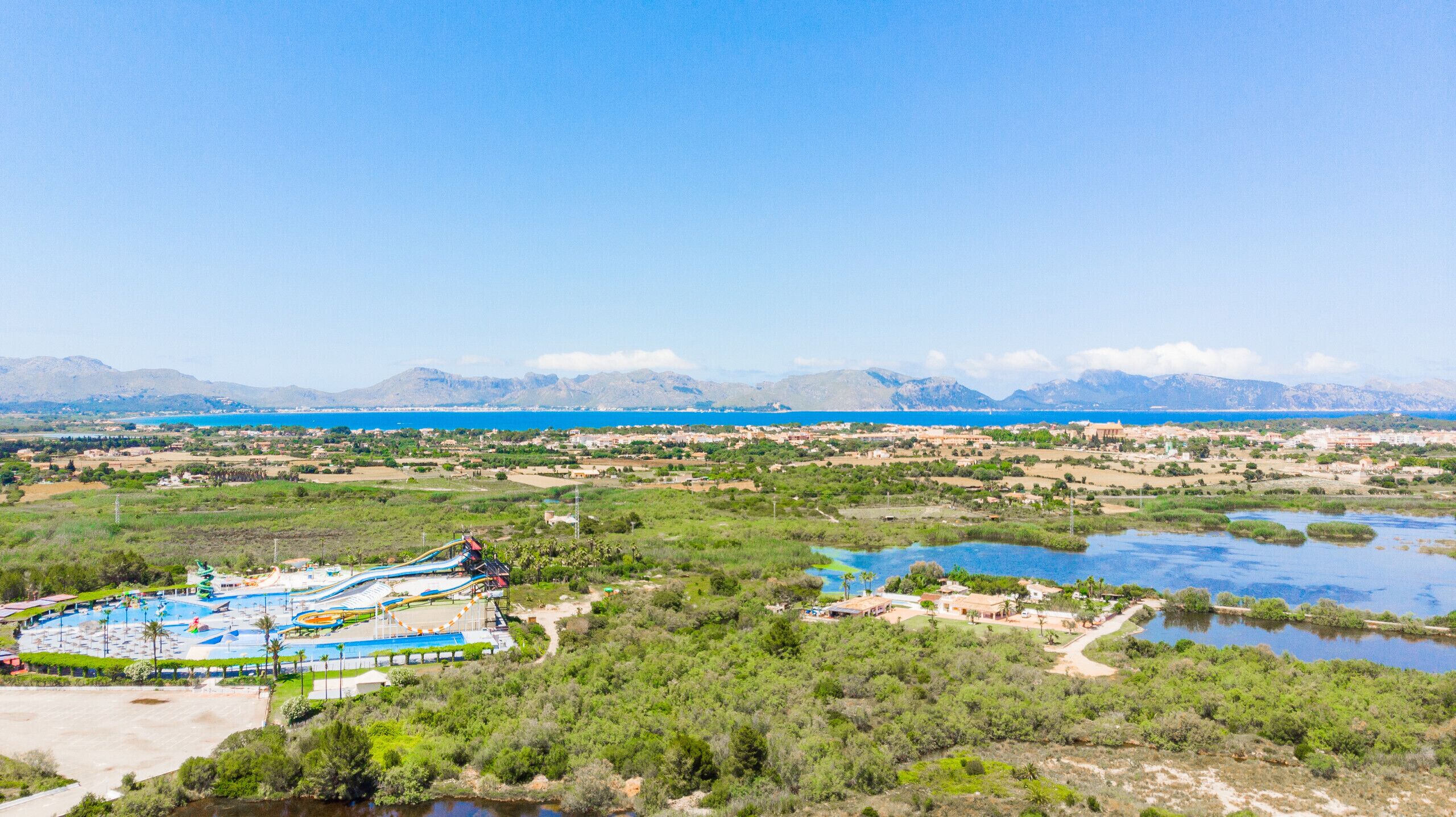 Ein Küstenresort mit Wasserpark, darunter ein Wellenbecken und Rutschen, umgeben von Bergen und einem klaren, blauen Meer.