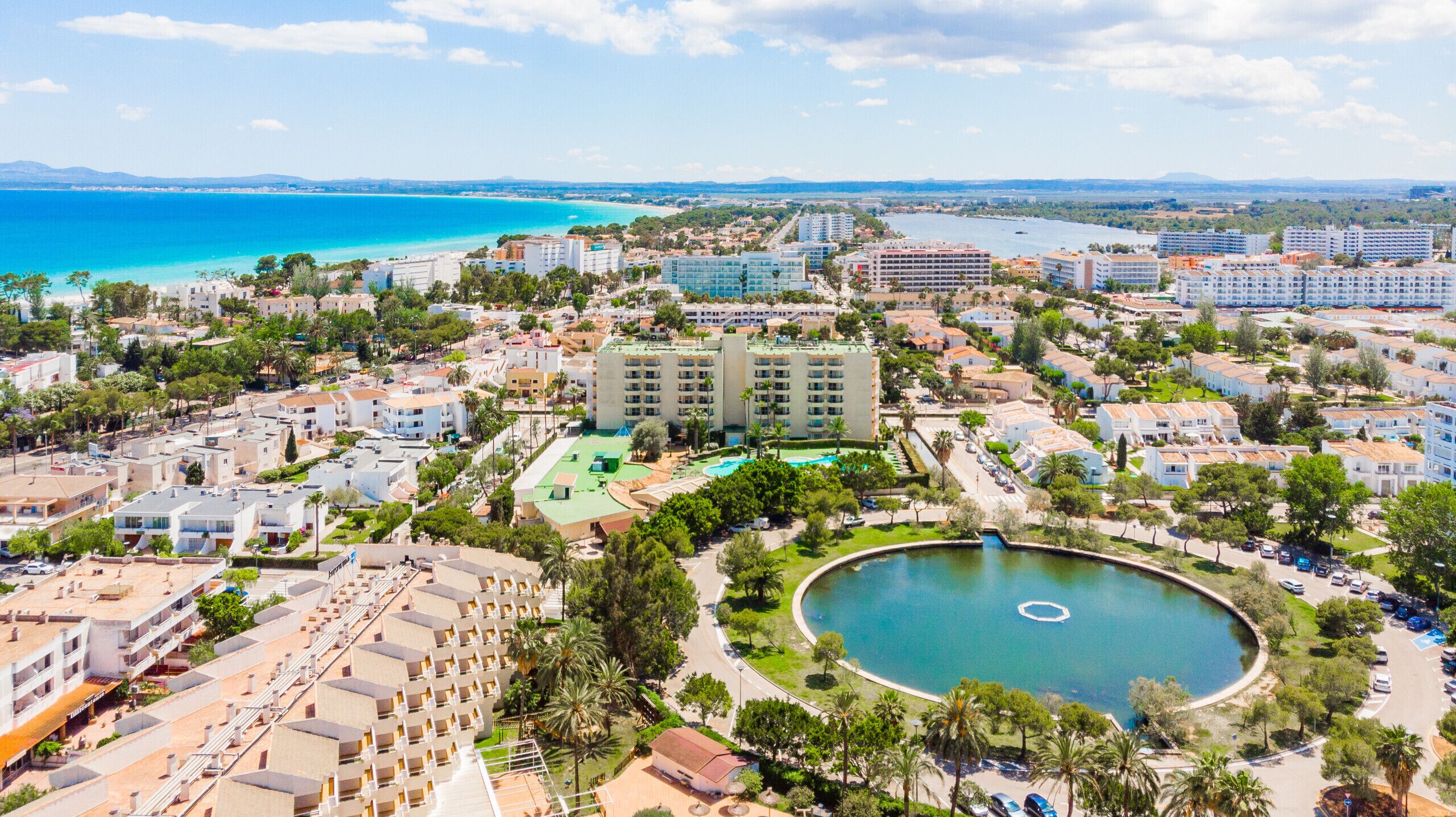 Vista aérea de uma área de resort costeira, com vários edifícios, uma piscina central e vista para o mar.