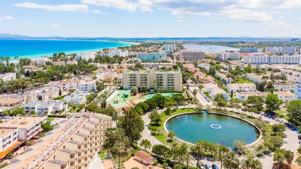 An aerial view of a coastal resort area with multiple buildings, a central swimming pool, and a view of the sea.