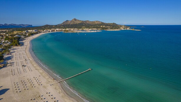 Vue aérienne d’une ville côtière avec une plage, une jetée et une montagne en arrière-plan.