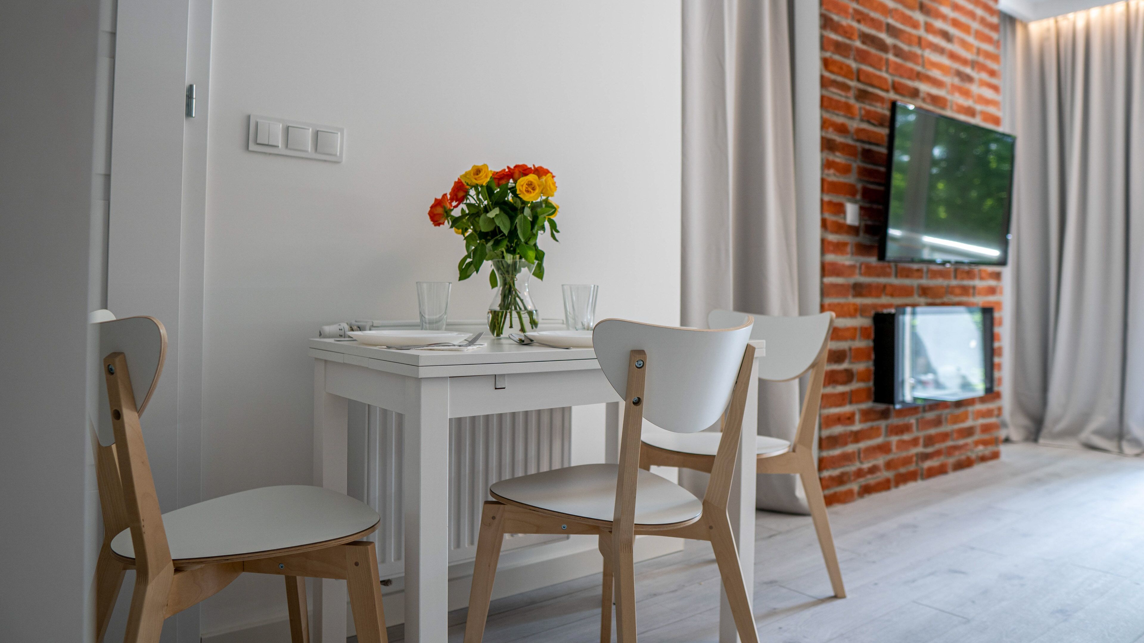 A dining area with a white table, two wooden chairs, a vase with flowers, and a TV mounted on a brick wall.