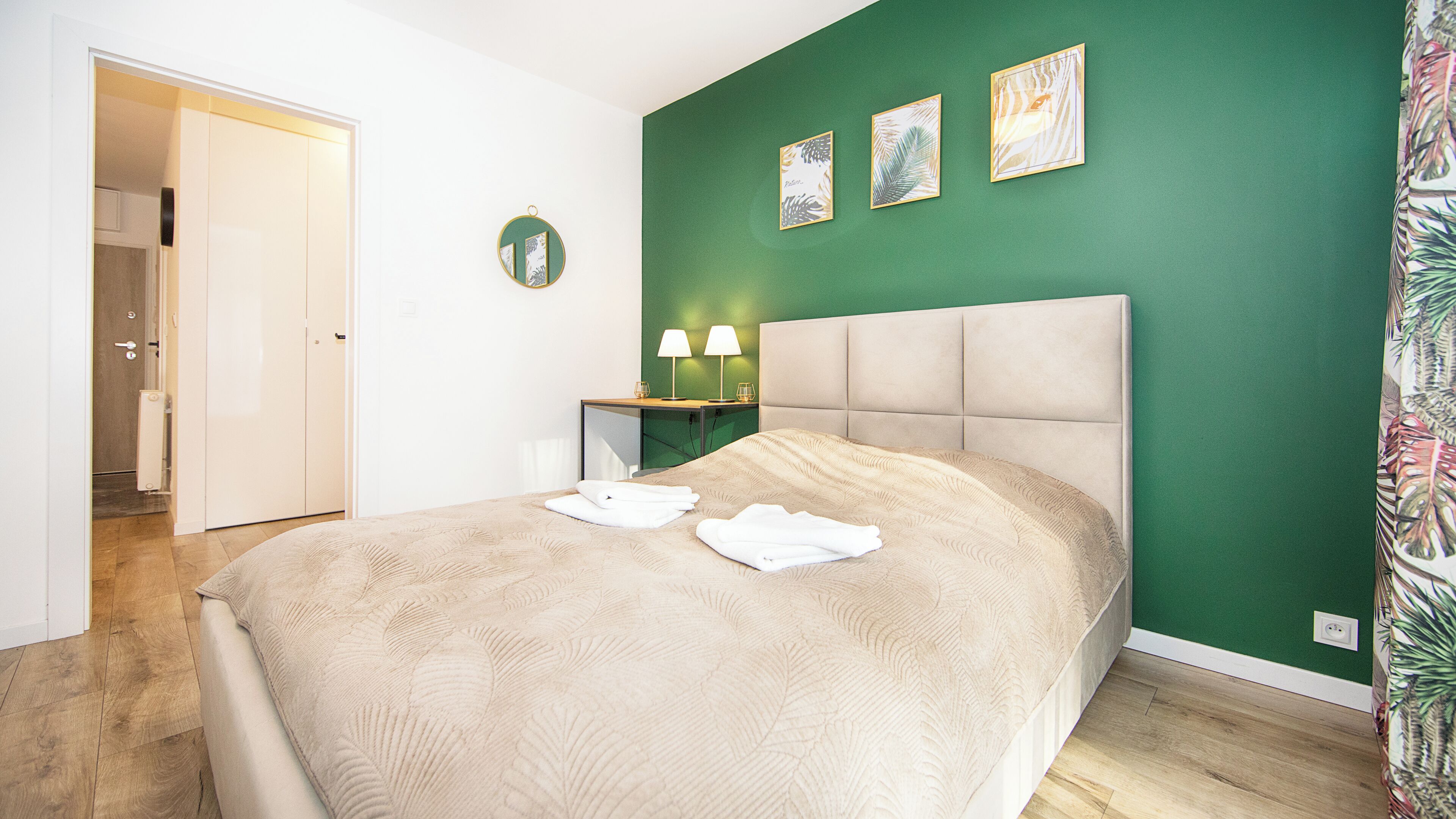 Una cama bien hecha con edredón color beige y toallas blancas, una mesita de noche con dos lámparas, y una pared verde con tres cuadros enmarcados.