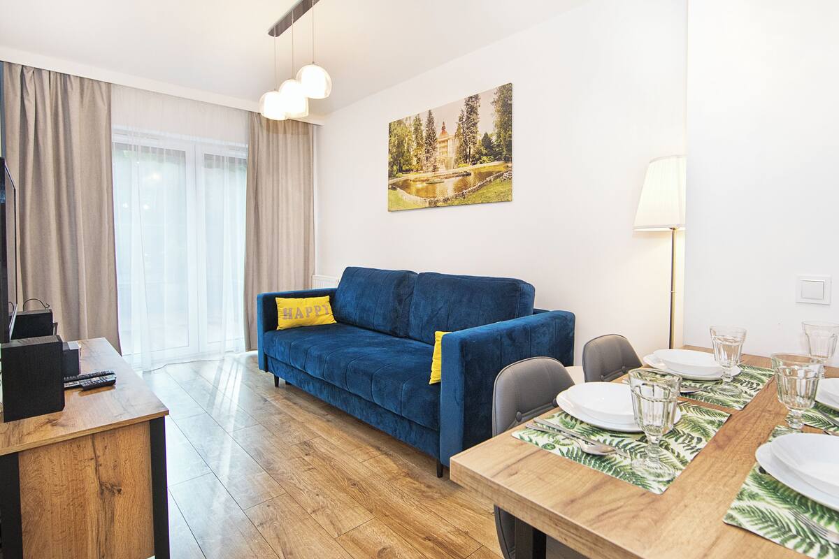 Una sala de estar moderna con un sofá azul, una mesa de comedor con platos y vasos, un cuadro en la pared y una lámpara.