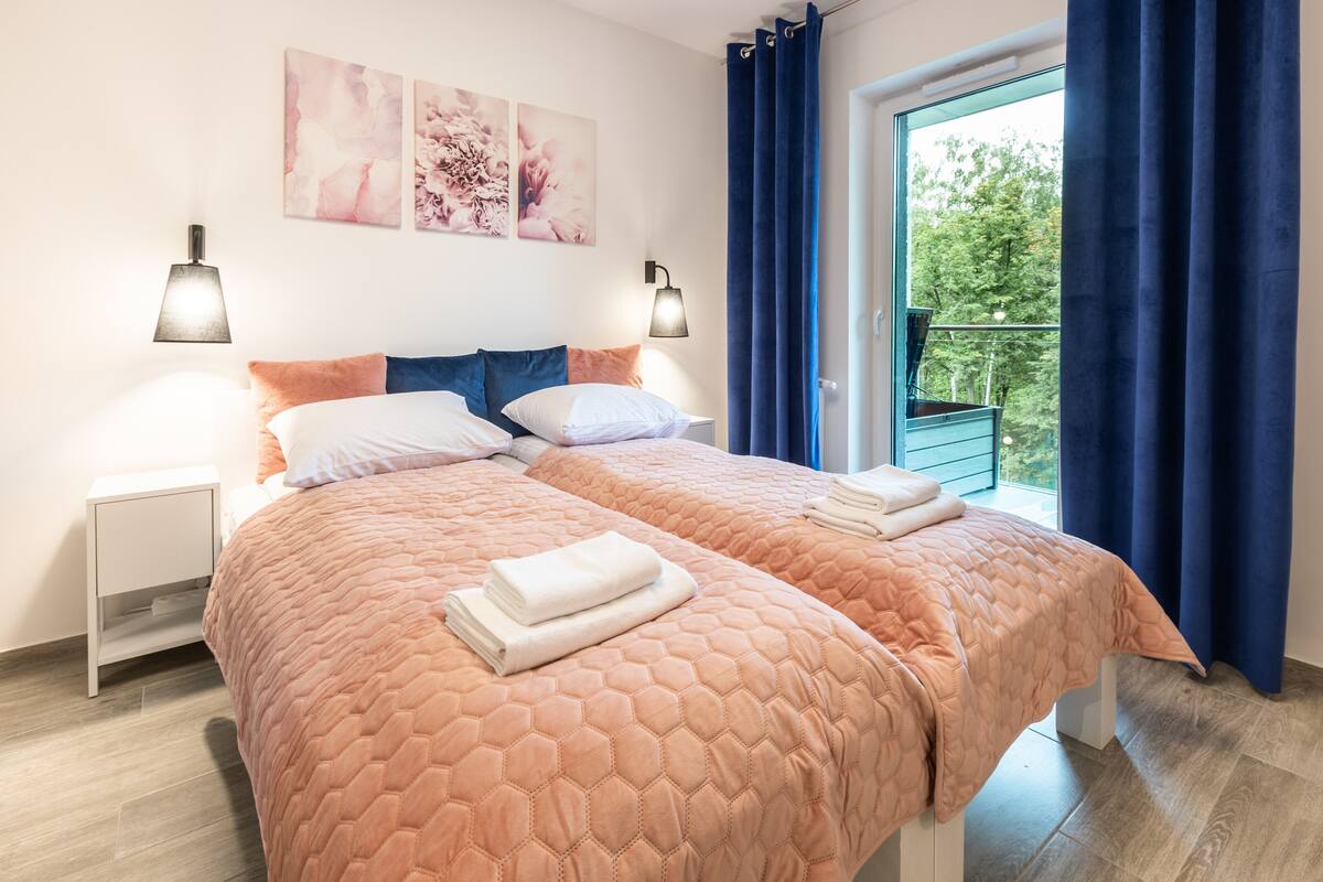 Una cama doble con colcha naranja acolchada, dos mesitas de noche con lámparas y una ventana con cortinas azules.