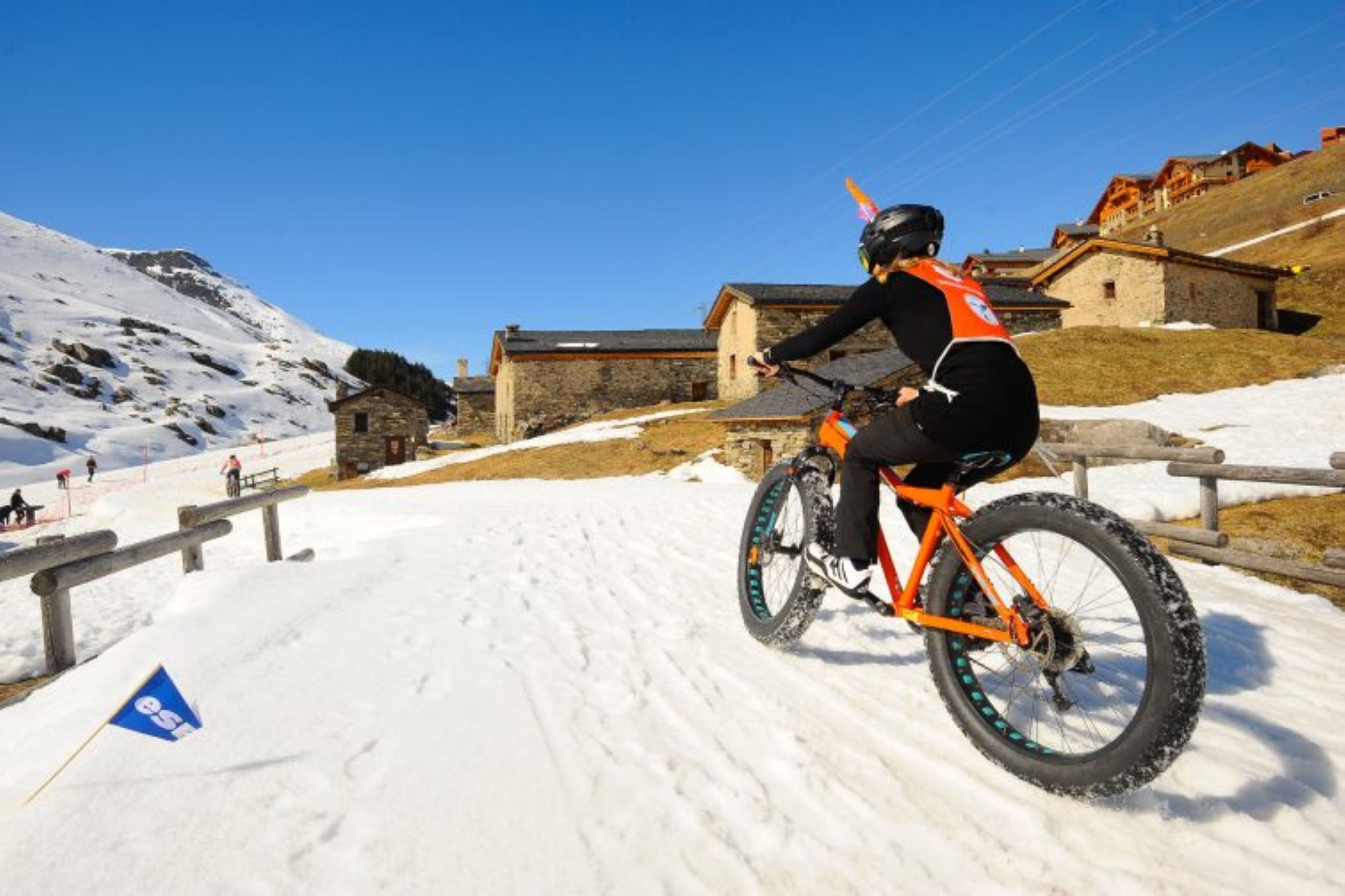 Uma pessoa anda de bicicleta de montanha numa trilha coberta de neve, com edifícios ao fundo.