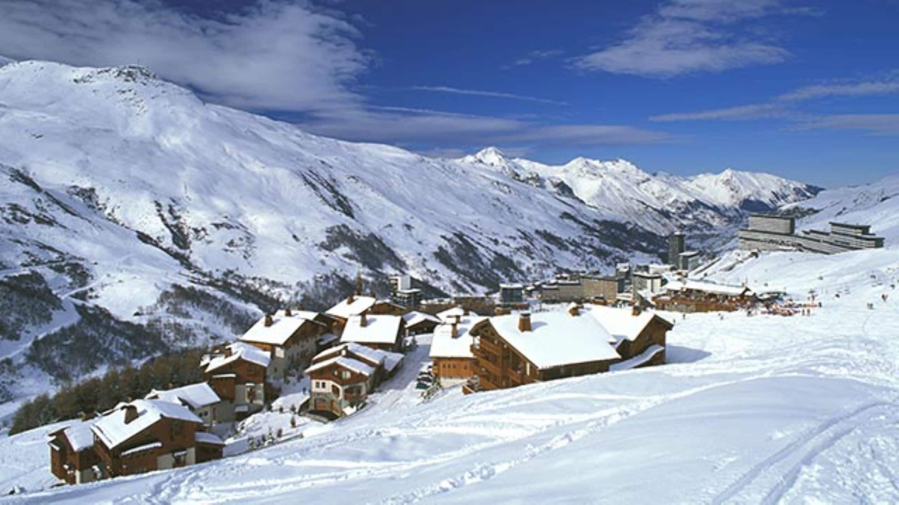 A snow-covered mountain village with wooden houses and a ski resort in the background.