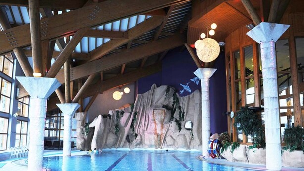 An indoor swimming pool with wooden beams, a rocky wall mural, and a seating area with a colorful chair.