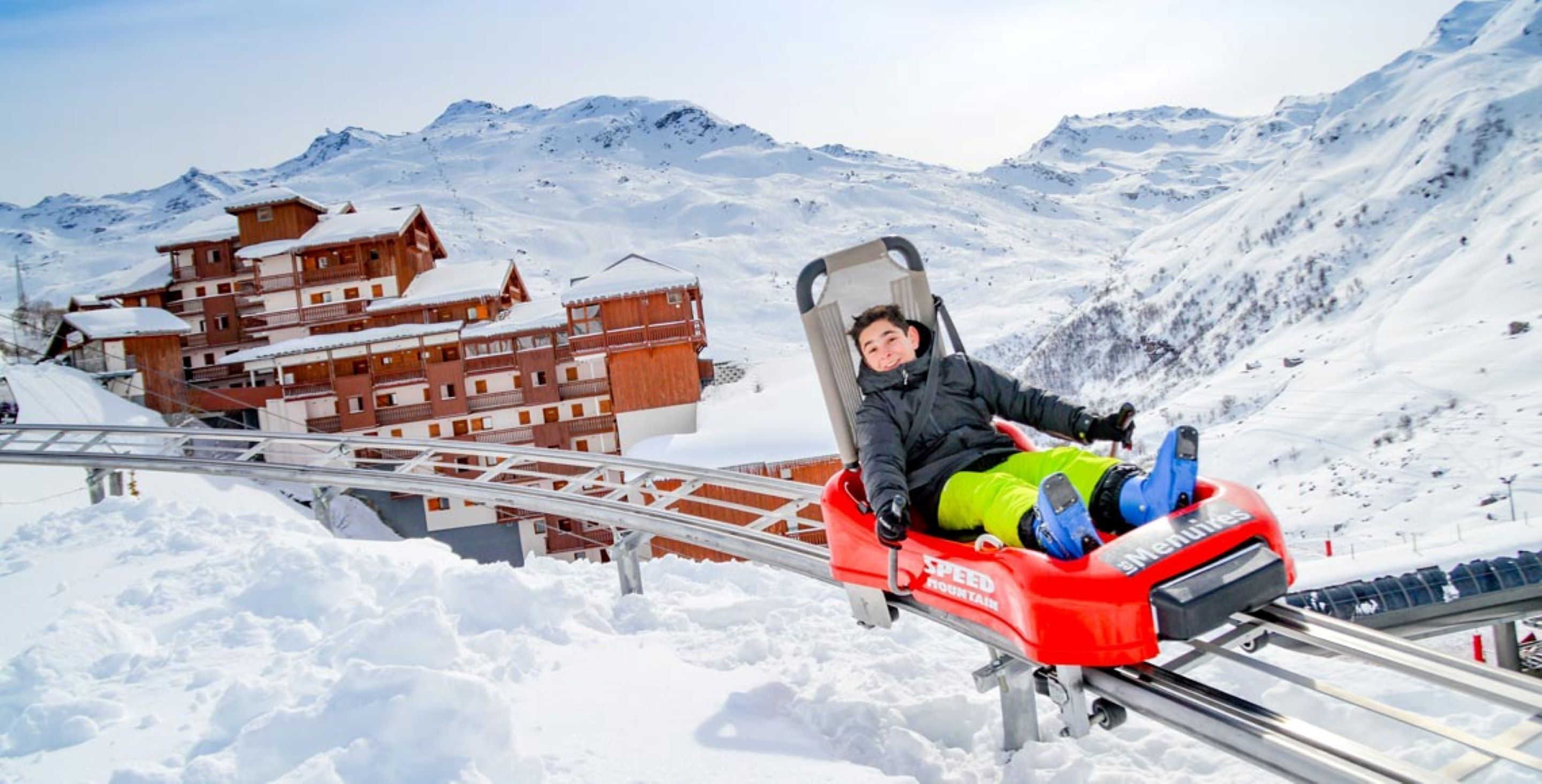 A person on a red sled, a wooden building, and snow-covered mountains.