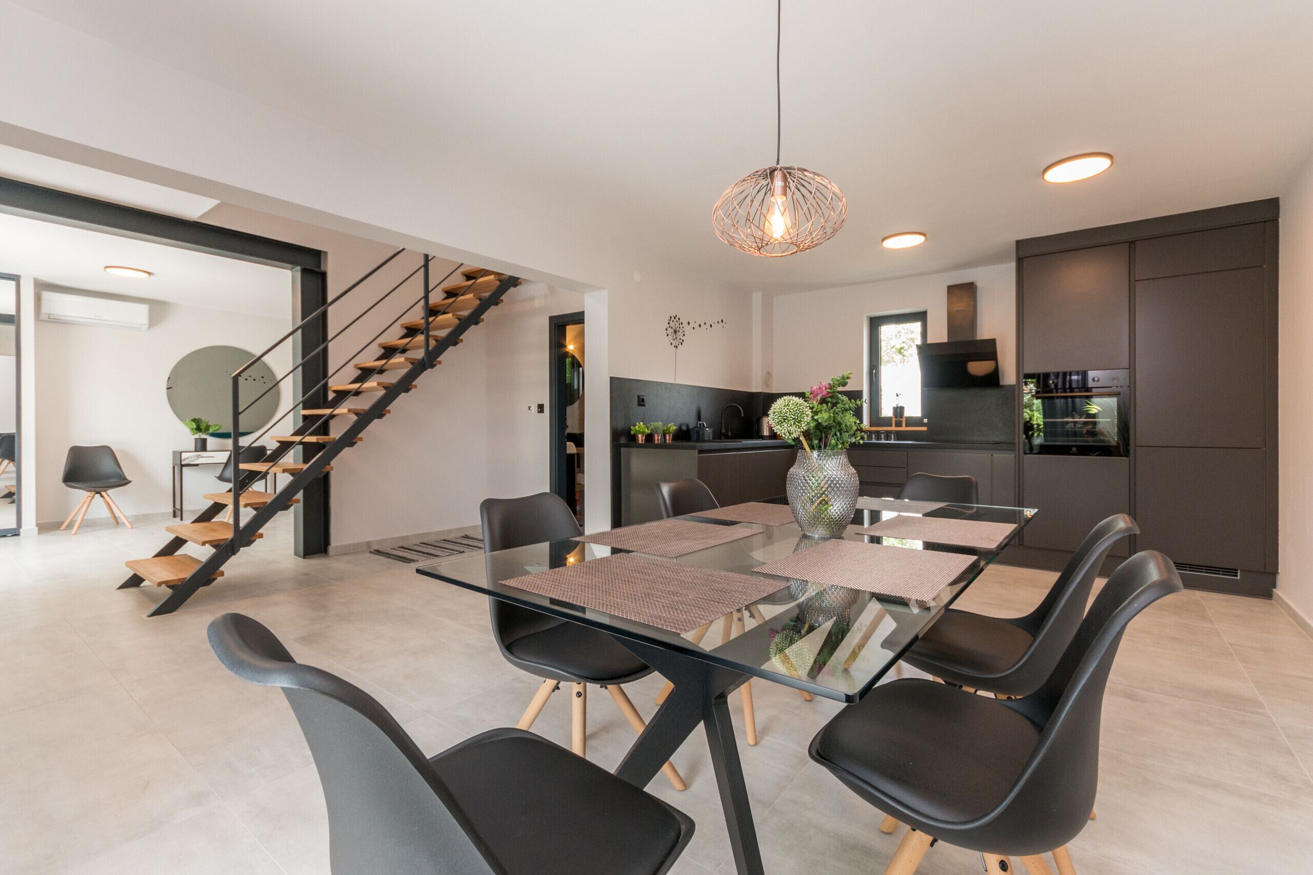 A modern kitchen with dark cabinets, a built-in oven, and a dining area with a glass table and black chairs.