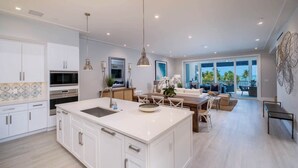 Une cuisine moderne avec des meubles blancs, un grand îlot central et un coin repas offrant une vue sur l’océan.