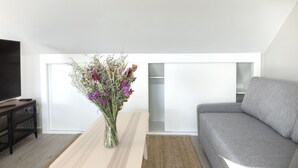 A modern living room with a grey sofa, a wooden coffee table with a vase of flowers, a flat-screen TV on a black stand, and a large window with a view of the sky.