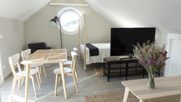 A modern, well-lit room with a dining table, chairs, a bed, a TV, and a vase of flowers.