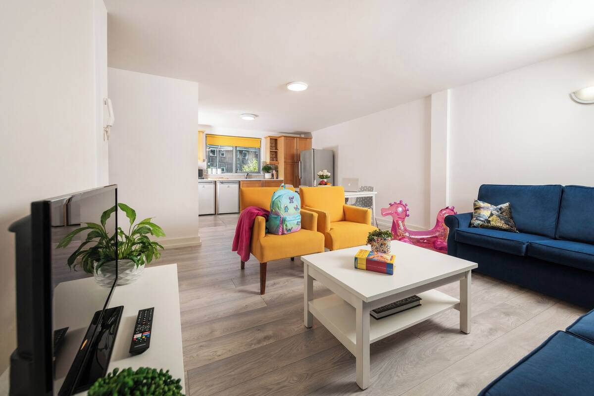 A modern living room with a white coffee table, a blue sofa, and a yellow armchair. A kitchen with yellow cabinets and a refrigerator is visible in the background.