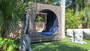 Un espacio de estar al aire libre con forma circular, que presenta una estructura tejida, rodeado de follaje y una estatua de león de piedra.