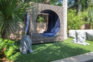 Un espacio de estar al aire libre con forma circular, que presenta una estructura tejida, rodeado de follaje y una estatua de león de piedra.
