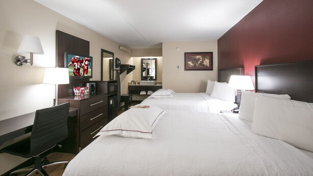 A hotel room with two beds, a desk, a chair, a TV, a mirror, and a red accent wall.