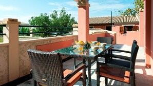 Mesa con cubierta de vidrio, sillas de mimbre y un frutero en un balcón con vistas a un edificio y árboles.