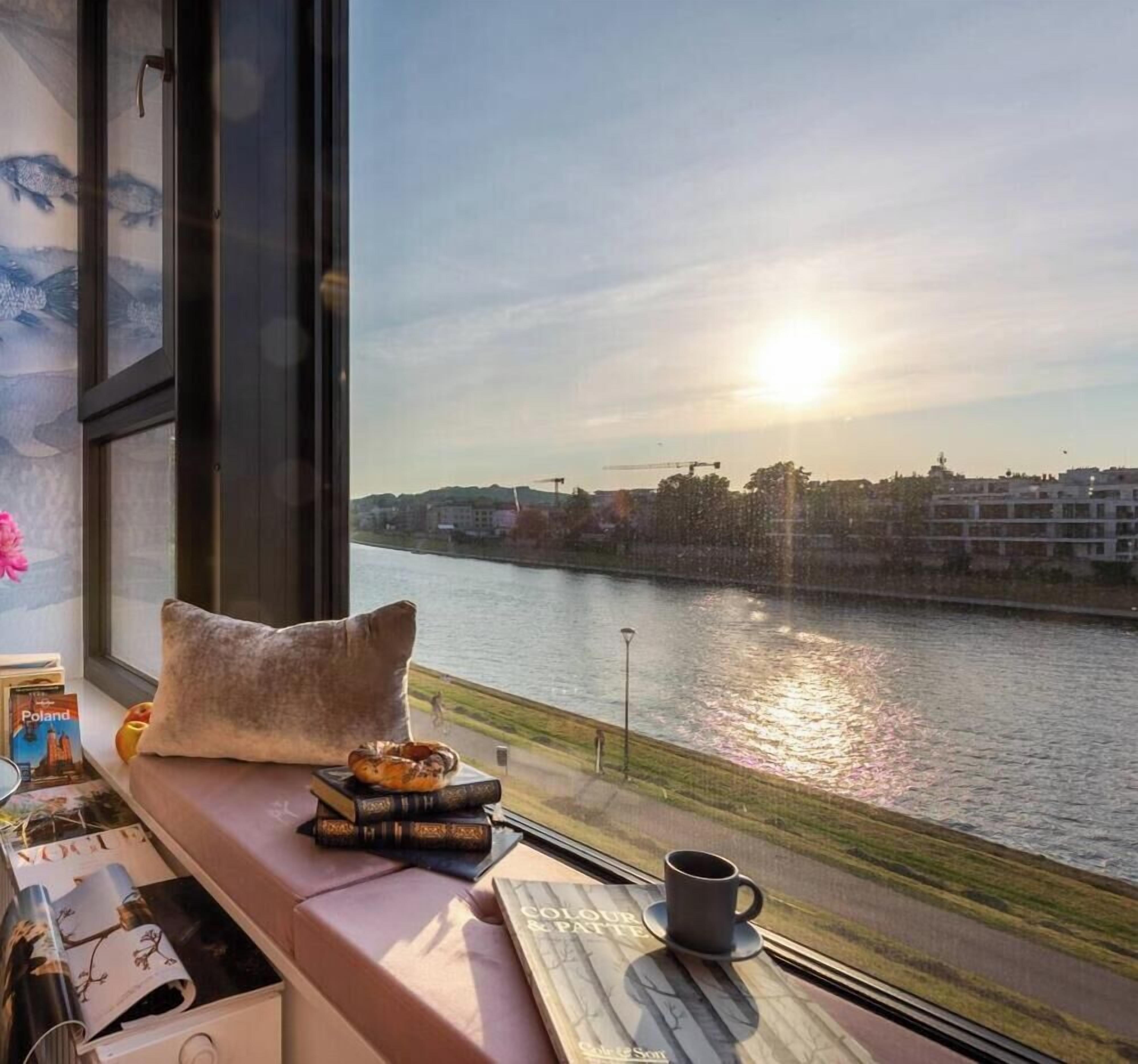 A room with a large window overlooking a river, a sofa with books and a mug, and a view of the cityscape.