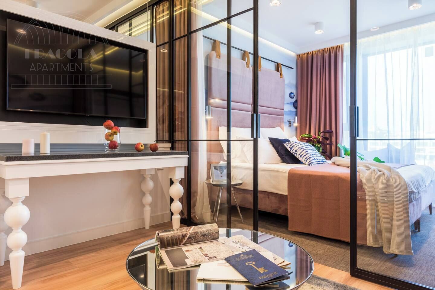 A modern hotel room with a large TV, a glass coffee table with magazines, a bed with a view, and a large window.