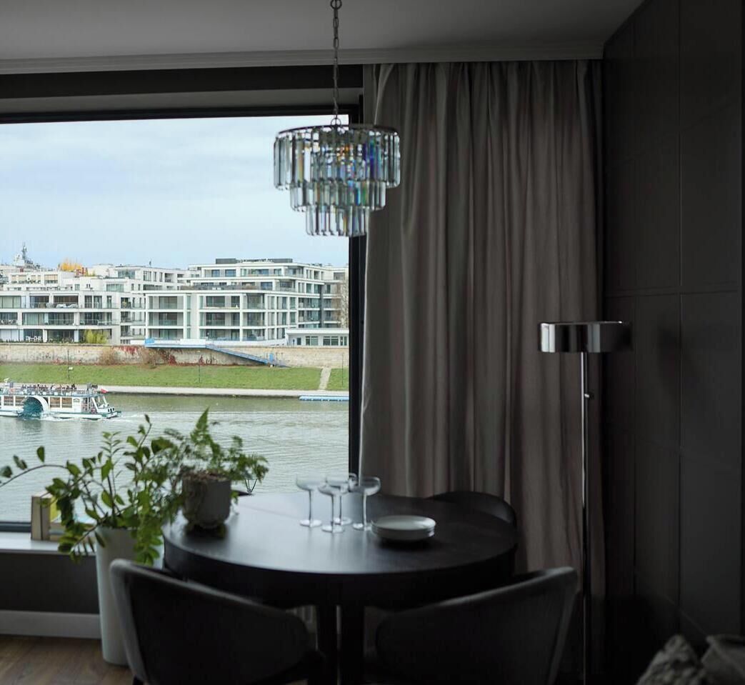 Una habitación moderna con vistas a un río y edificios, una mesa de comedor redonda con sillas, una planta en maceta y una lámpara de pie alta.