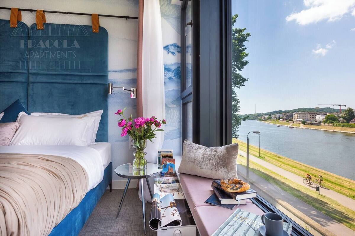 Habitación de hotel moderna con una cama grande, una mesita con un florero y vistas a un río y al paisaje urbano.