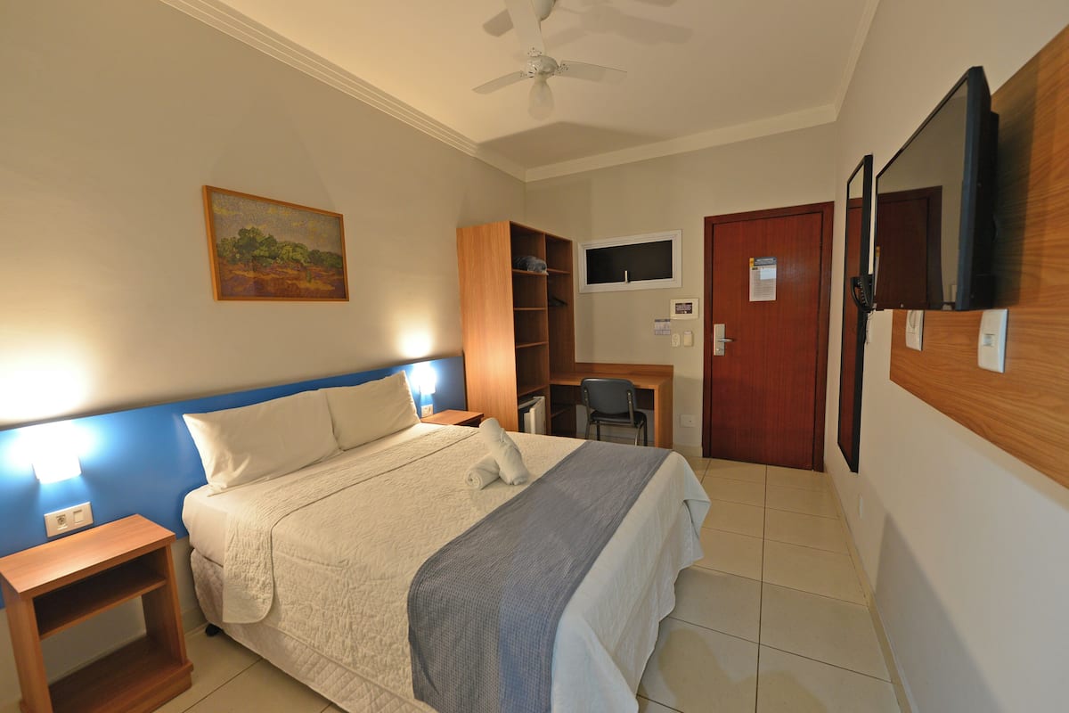 Ein Hotelzimmer mit Bett, Schreibtisch, Stuhl, Deckenventilator, einem Bild und einem Fernseher an der Wand.