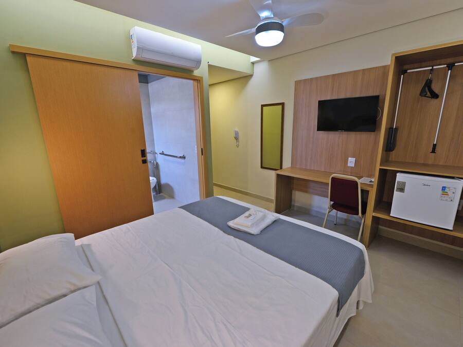 Quarto de hotel com cama, escrivaninha, cadeira, guarda-roupa, televisão e frigorífico.