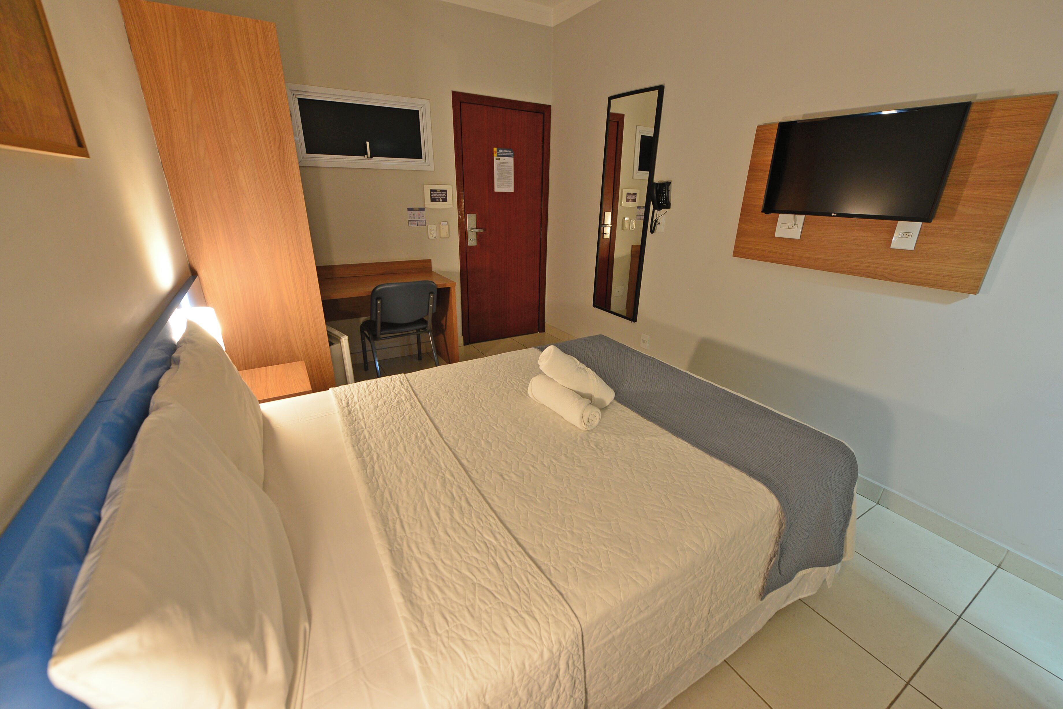 Ein Hotelzimmer mit Bett, Schreibtisch, Stuhl, Tür und einem an der Wand montierten Fernseher.